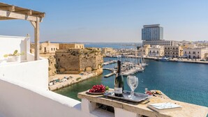 Una terraza frente al mar con vistas a un antiguo fuerte de piedra, un puerto deportivo con barcos y un edificio moderno al fondo.