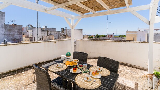Une terrasse sur le toit, avec une table dressée pour le dîner, entourée de chaises en osier, sous une pergola en bois.
