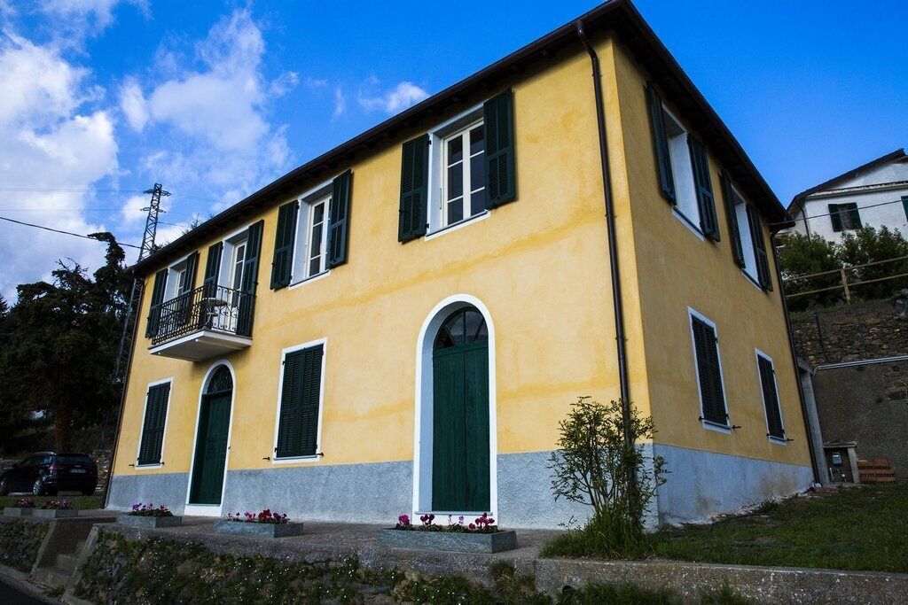 A yellow building with green shutters, a balcony, and a small garden area.