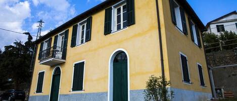 A yellow building with green shutters, a balcony, and a small garden area.