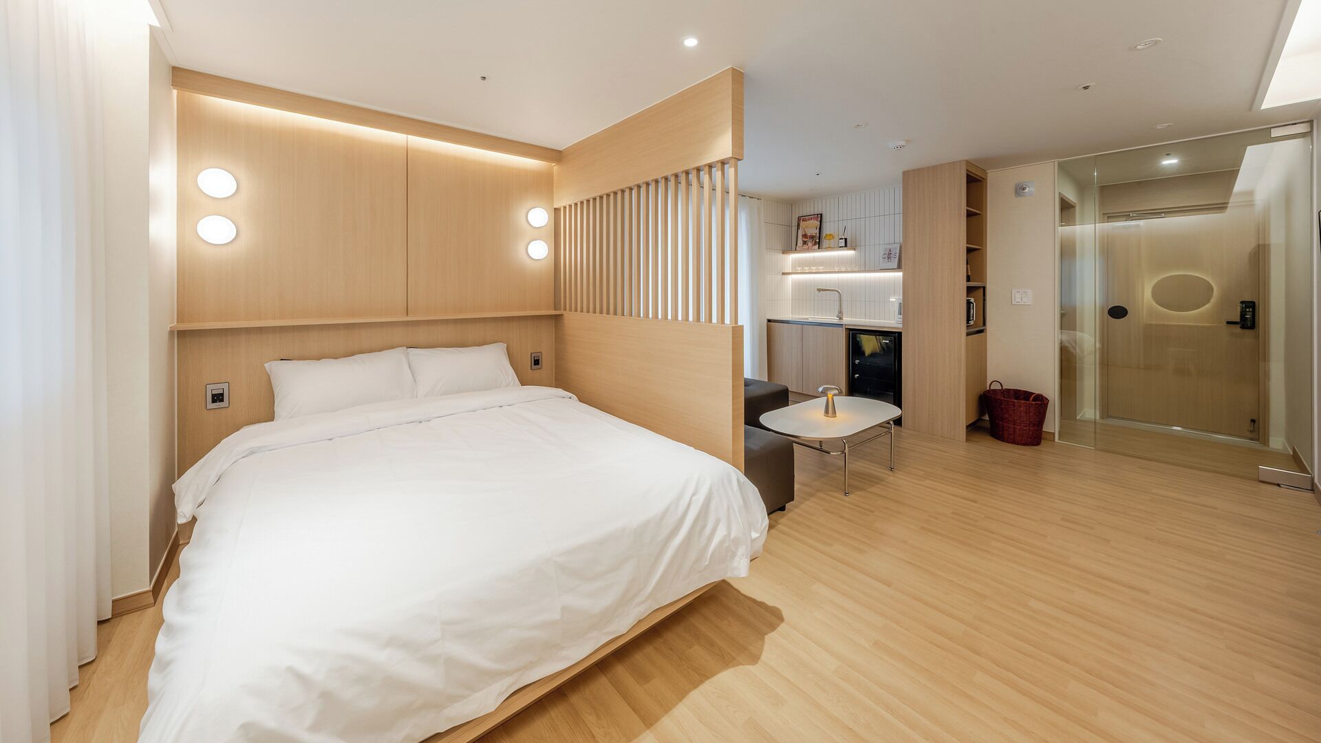 Una habitación de hotel moderna con una cama grande, una mesita pequeña, una cocina integrada y un box de ducha de vidrio.