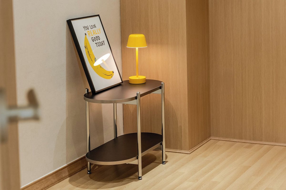 Eine Ecke mit einem gerahmten Motivationsspruch, einem kleinen Tisch mit einer gelben Lampe und einem Holzboden.