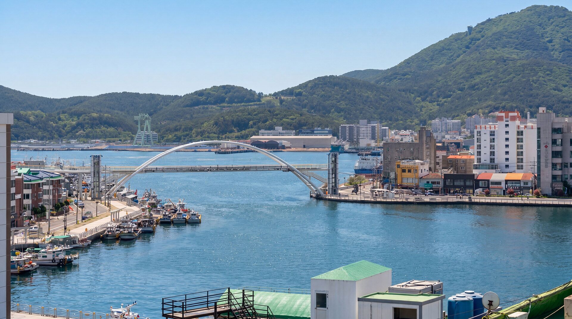 Un paisaje urbano costero con un puente destacado, un puerto deportivo con barcos y edificios industriales.