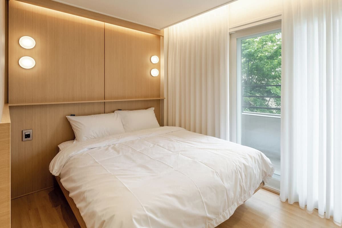 Ein modernes Schlafzimmer mit einem hölzernen Kopfende, einem großen Bett und Blick auf Grün hinter durchsichtigen Vorhängen.