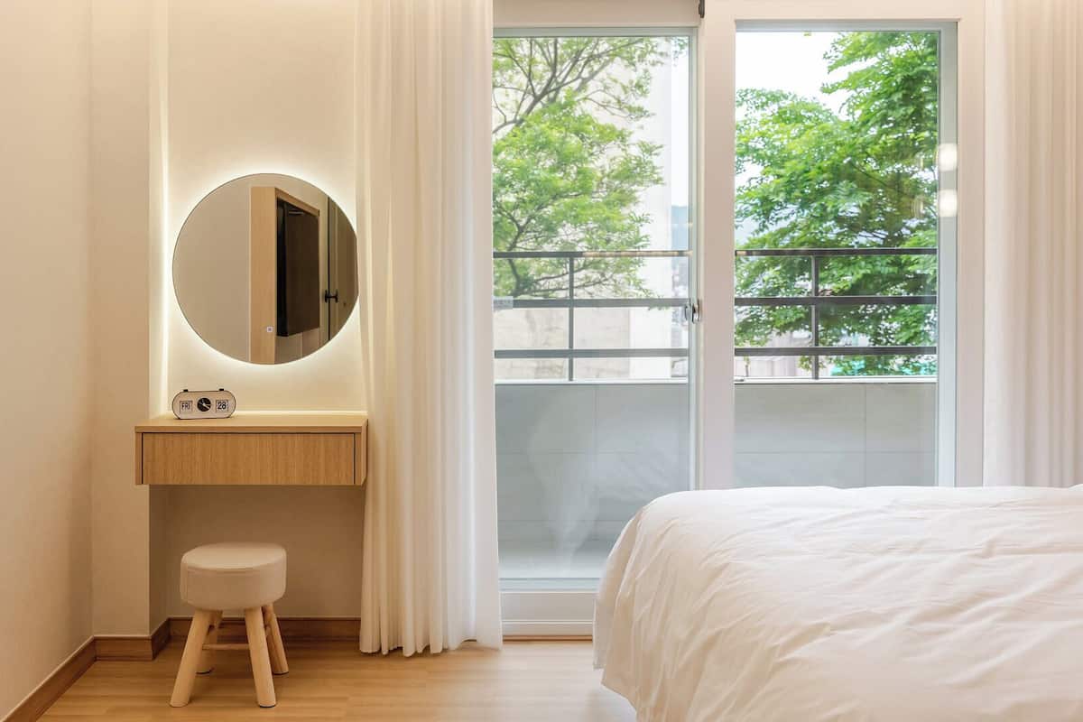 Ein Schlafzimmer mit Bett, Nachttisch mit Uhr, Spiegel und Blick auf Bäume durch eine Schiebetür.