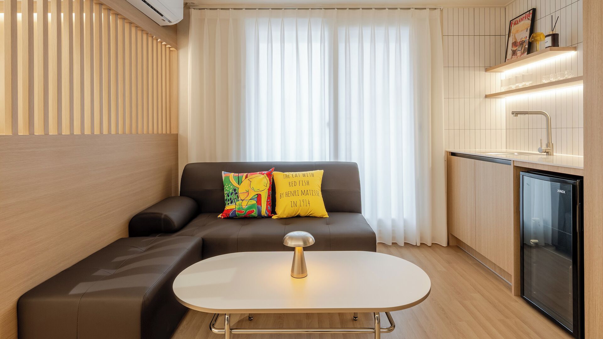 Un espacio habitable compacto con sofá, una mesita, kitchenette y una ventana con cortinas.