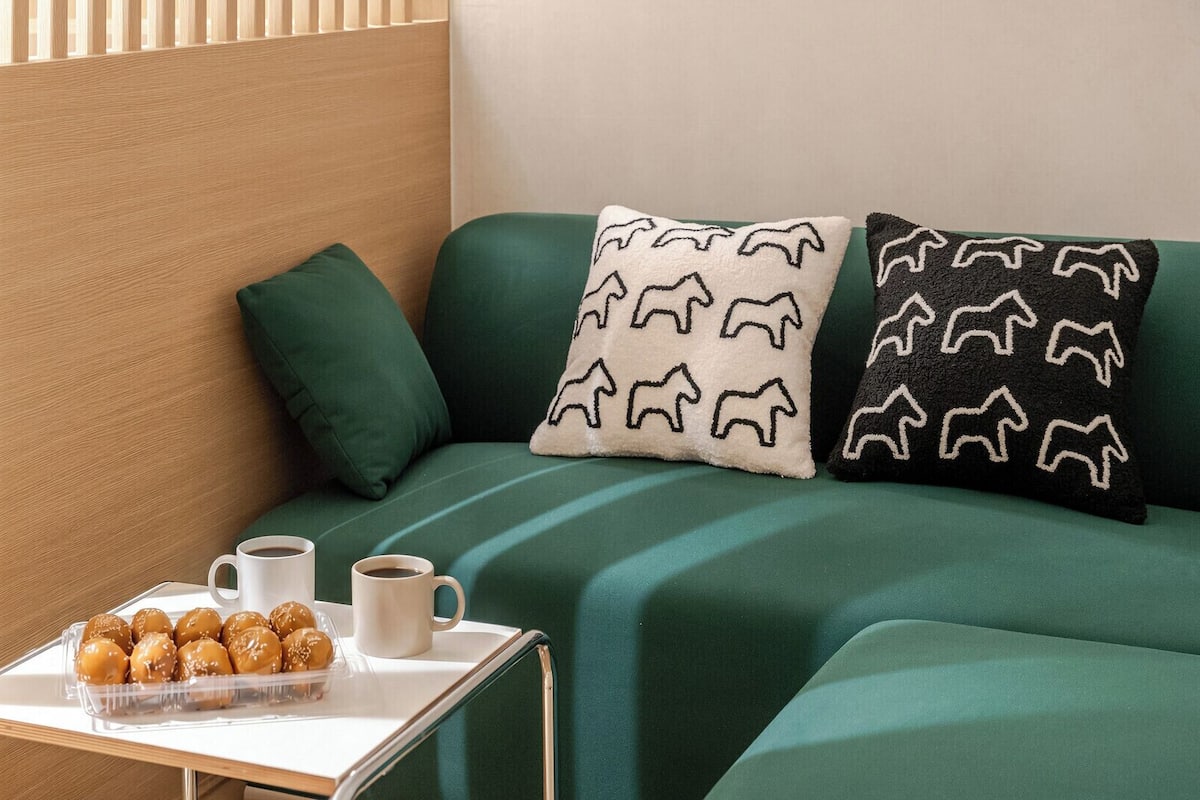 Ein Zimmer mit einem hölzernen Kopfende, einem grünen Stoffsofa, zwei gemusterten Kissen, einem kleinen Tisch mit einer Snackplatte und zwei Tassen sowie einem Fenster mit Jalousien.