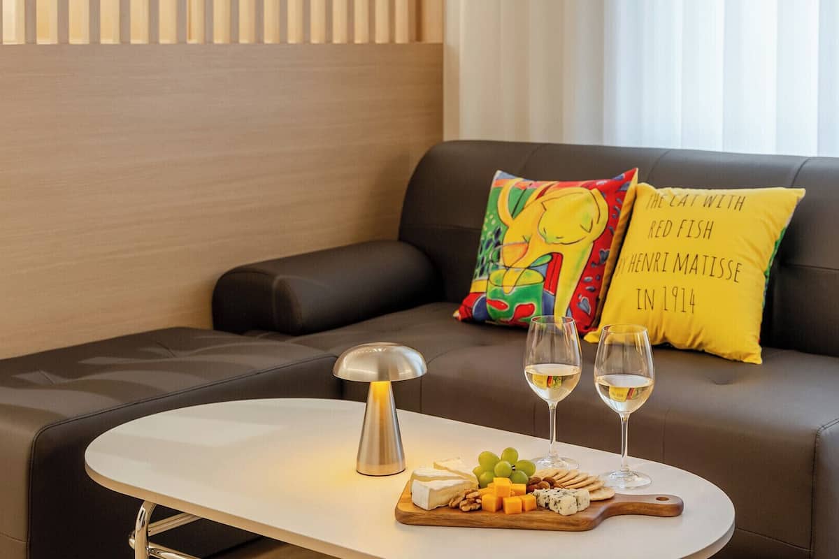 Ein modernes Wohnzimmer mit einer Couch, einem kleinen Tisch mit Lampe, Gläsern Weißwein und einer Käseplatte mit verschiedenen Käsesorten und Crackern.