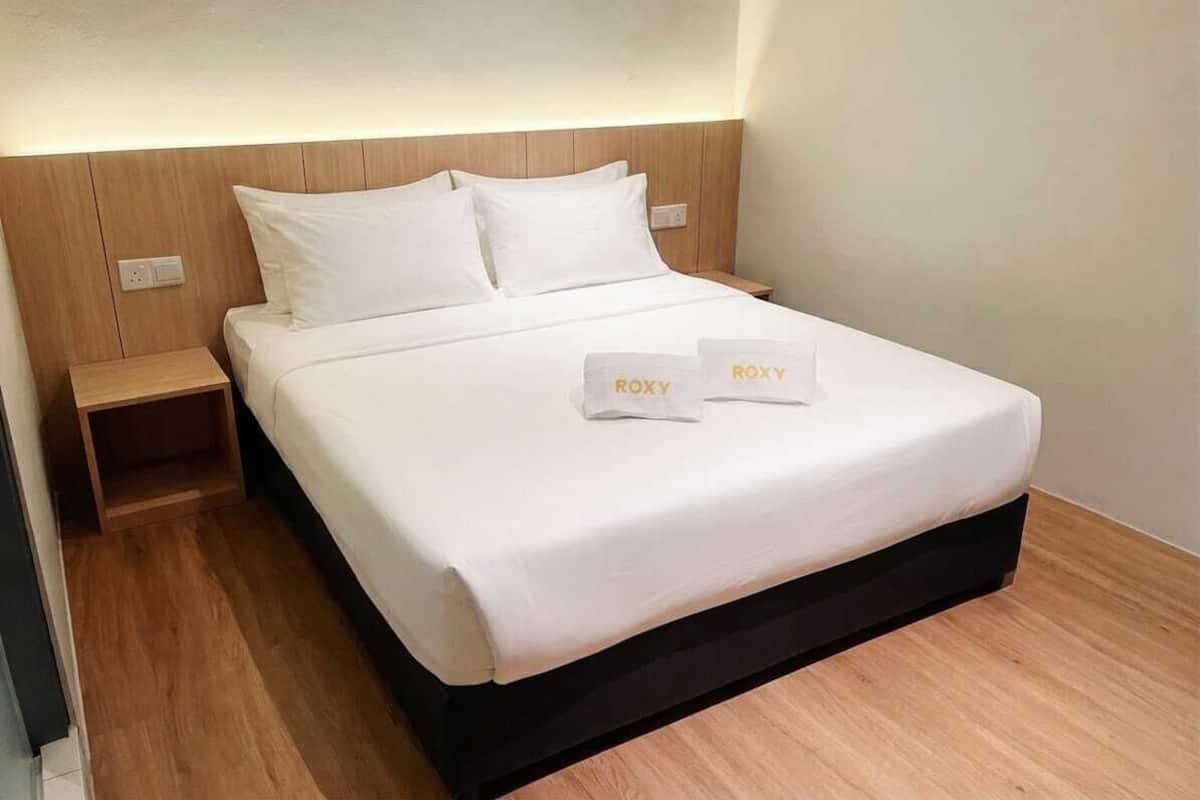 Una cama bien tendida con sábanas blancas y dos almohadas, un cabecero de madera y una mesita de noche con toma de corriente.