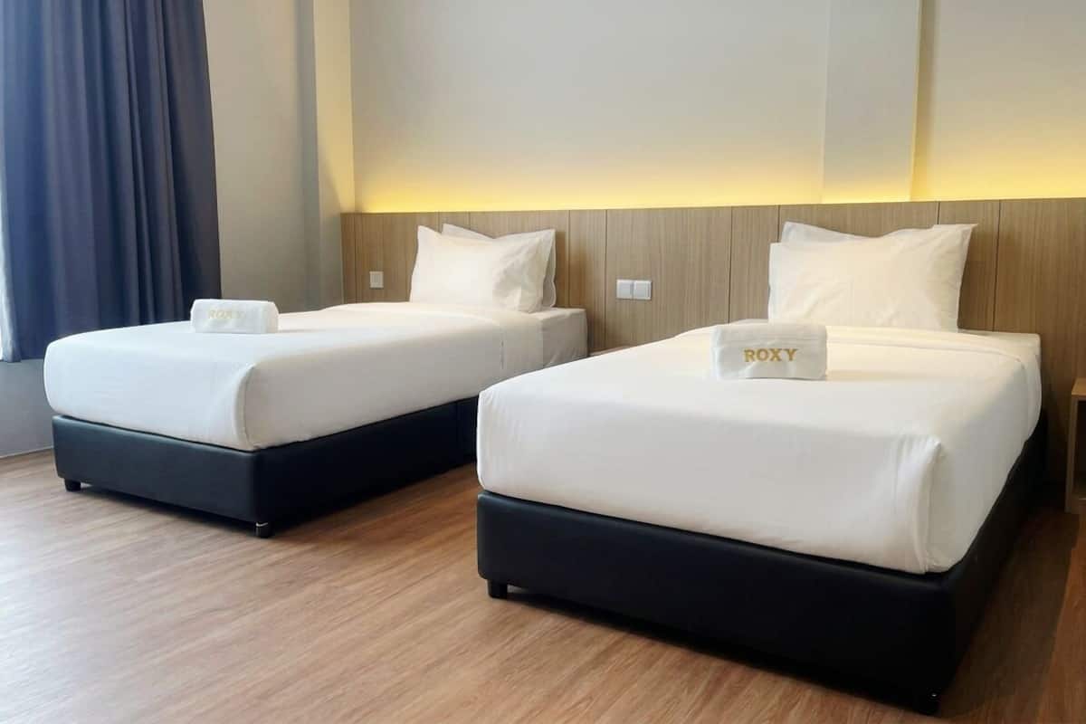 Dos camas con ropa de cama blanca y una etiqueta de almohada 'ROXY', un cabecero de madera y una ventana con cortinas.