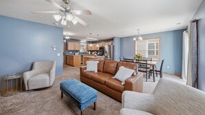 Ein Wohnzimmer mit braunem Ledersofa, zwei Sesseln, einem blauen Hocker, einem Deckenventilator und einer Küche im Hintergrund.