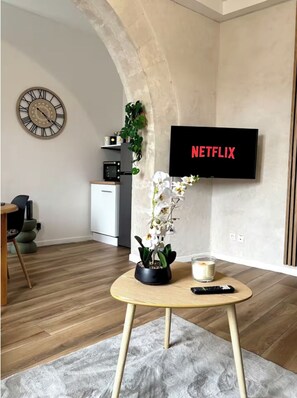Olohuoneessa on puulattia, pyöreä pöytä, jonka päällä on ruukkuorkidea, kaukosäädin, kynttilä ja seinälle asennettu televisio, jonka näytöllä näkyy teksti ”NETFLIX”.