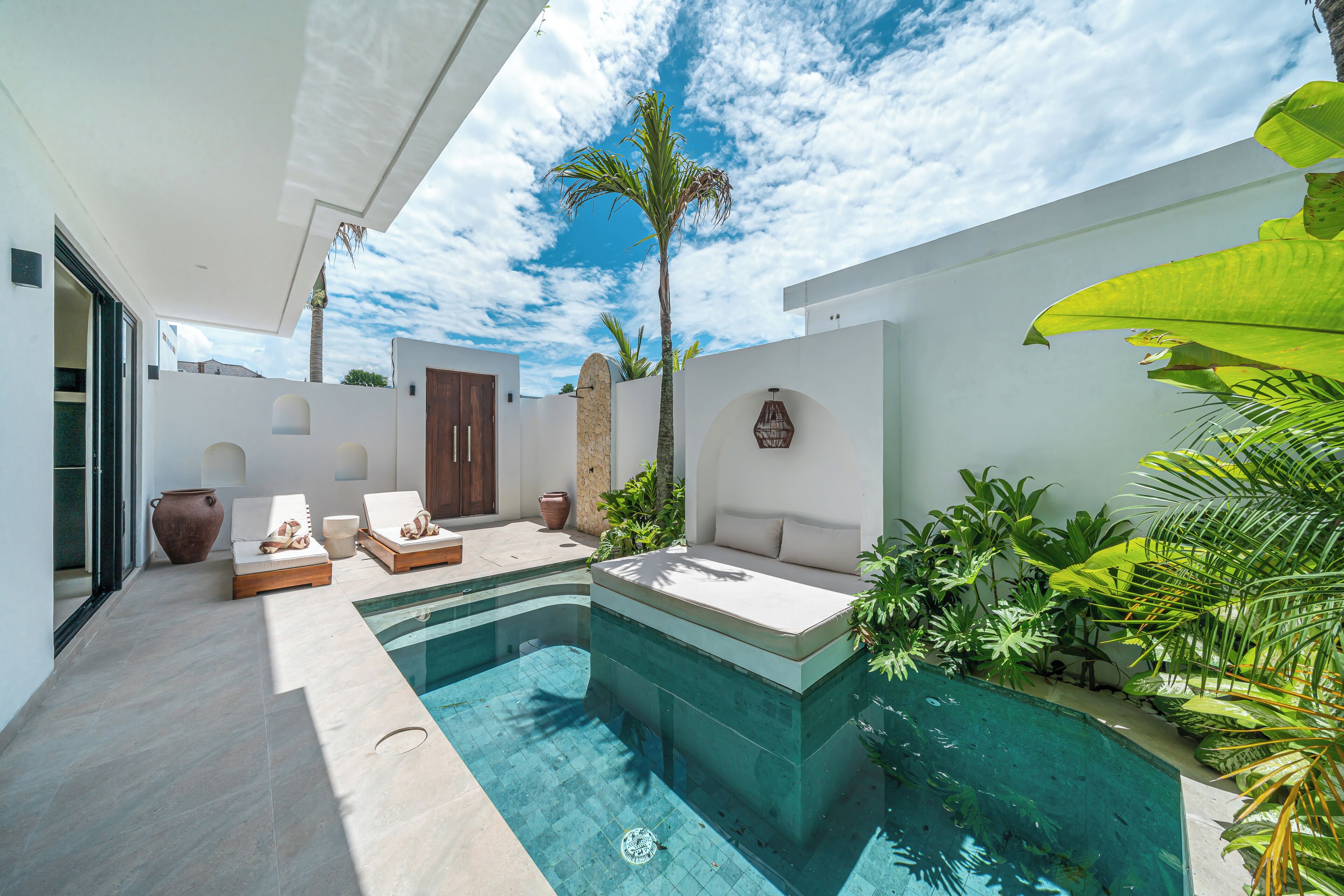 A modern outdoor pool area with a built-in seating area, surrounded by plants and a clear sky.