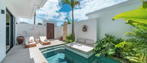 A modern outdoor pool area with a built-in seating area, surrounded by plants and a clear sky.