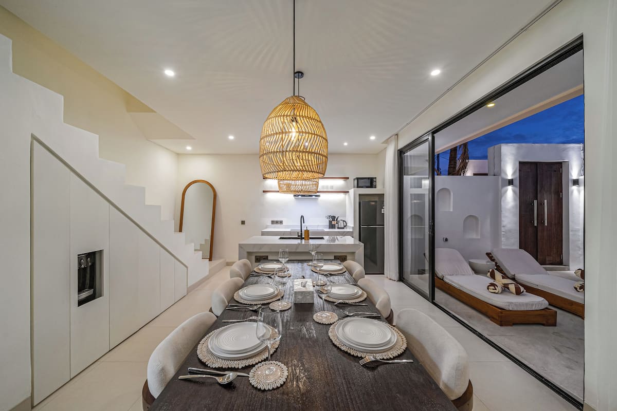 Un espace repas moderne avec une longue table dressée pour six personnes, une cuisine en arrière-plan et une cloison en verre donnant sur un coin salon.
