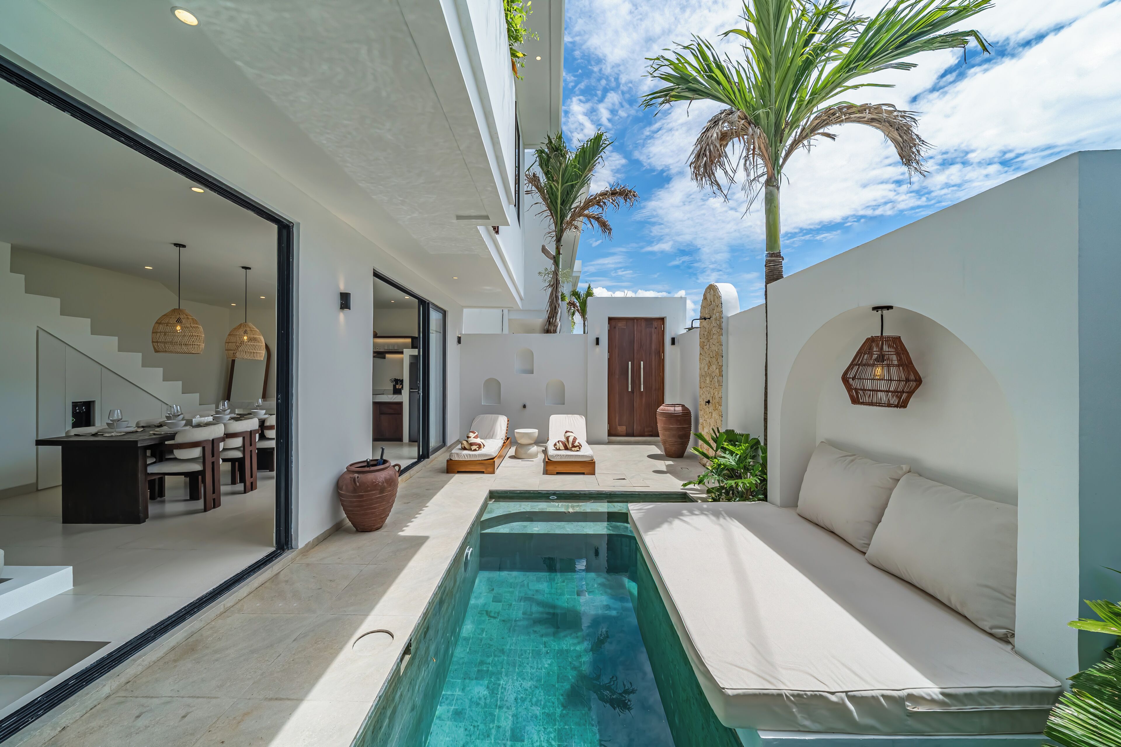 Uma área de piscina moderna com um sofá, espreguiçadeiras e uma sala de jantar visíveis através de uma porta de vidro.