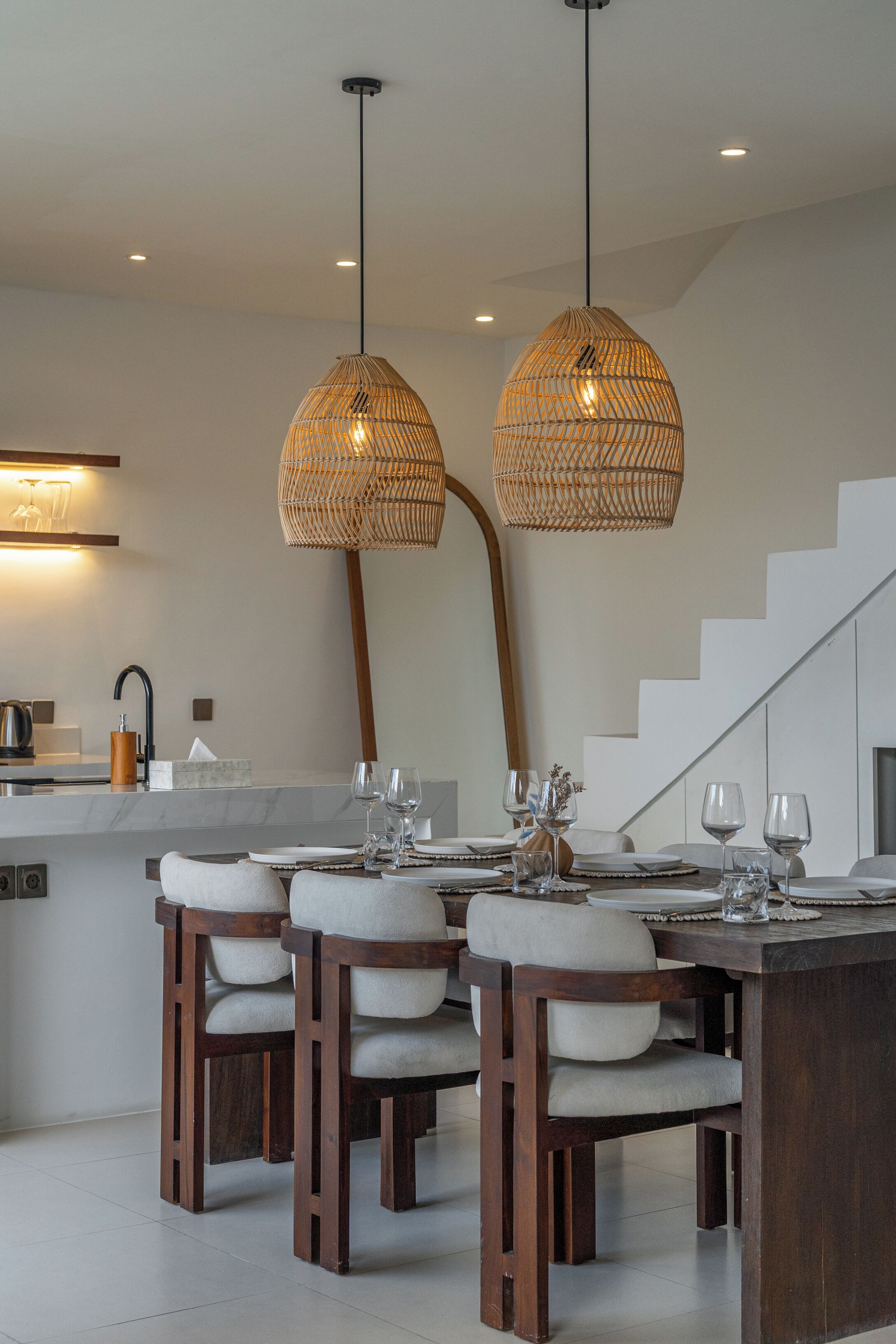 A modern kitchen with a dining area, featuring a central island with a built-in stove, wooden chairs, and pendant lighting.