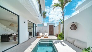 Un área moderna de piscina al aire libre con un sofá, sillones de descanso y una escalera que conduce a un espacio interior.