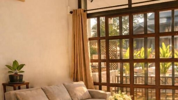 Una sala de estar con sofá, reposapiés y una mesa de centro, una planta en maceta y un amplio ventanal con cortinas.