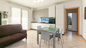 A modern kitchen with white cabinets, a glass dining table, and a brown sofa. - Antenore [relaxation with swimming pool] (Lido di Jesolo)