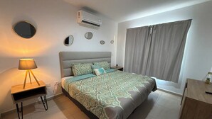 Une chambre à coucher comprenant un lit, une table de chevet, une lampe, un appareil de climatisation et une fenêtre avec des rideaux.
