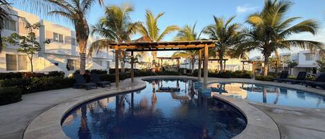A swimming pool with lounge chairs, palm trees, and modern buildings in the background.