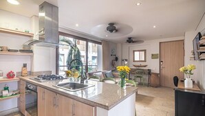 A modern kitchen with a central island, a stove, and a sink. There is a dining area with a table and chairs, and a view of the outside through large windows. - Designer apartment with pool and private balcony (Santa Marta)