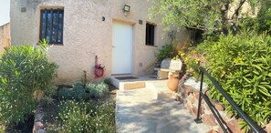 Ein Gebäude mit einer Fassade aus Stein, einer weißen Tür und einem kleinen Garten mit verschiedenen Pflanzen und einem roten Gartenschlauch.