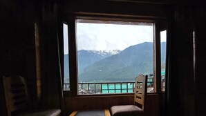 A room with a large window offering a view of mountains and a body of water. There are two wooden chairs and a small table. - The Park Green Resorts (Manali)