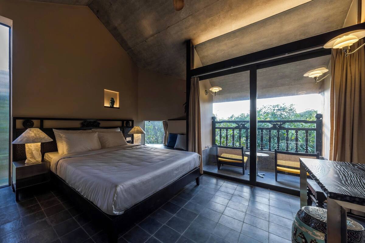 Un dormitorio con cama, lámparas de noche, ventilador de techo, un balcón con mesa y sillas, y vista a zonas verdes.