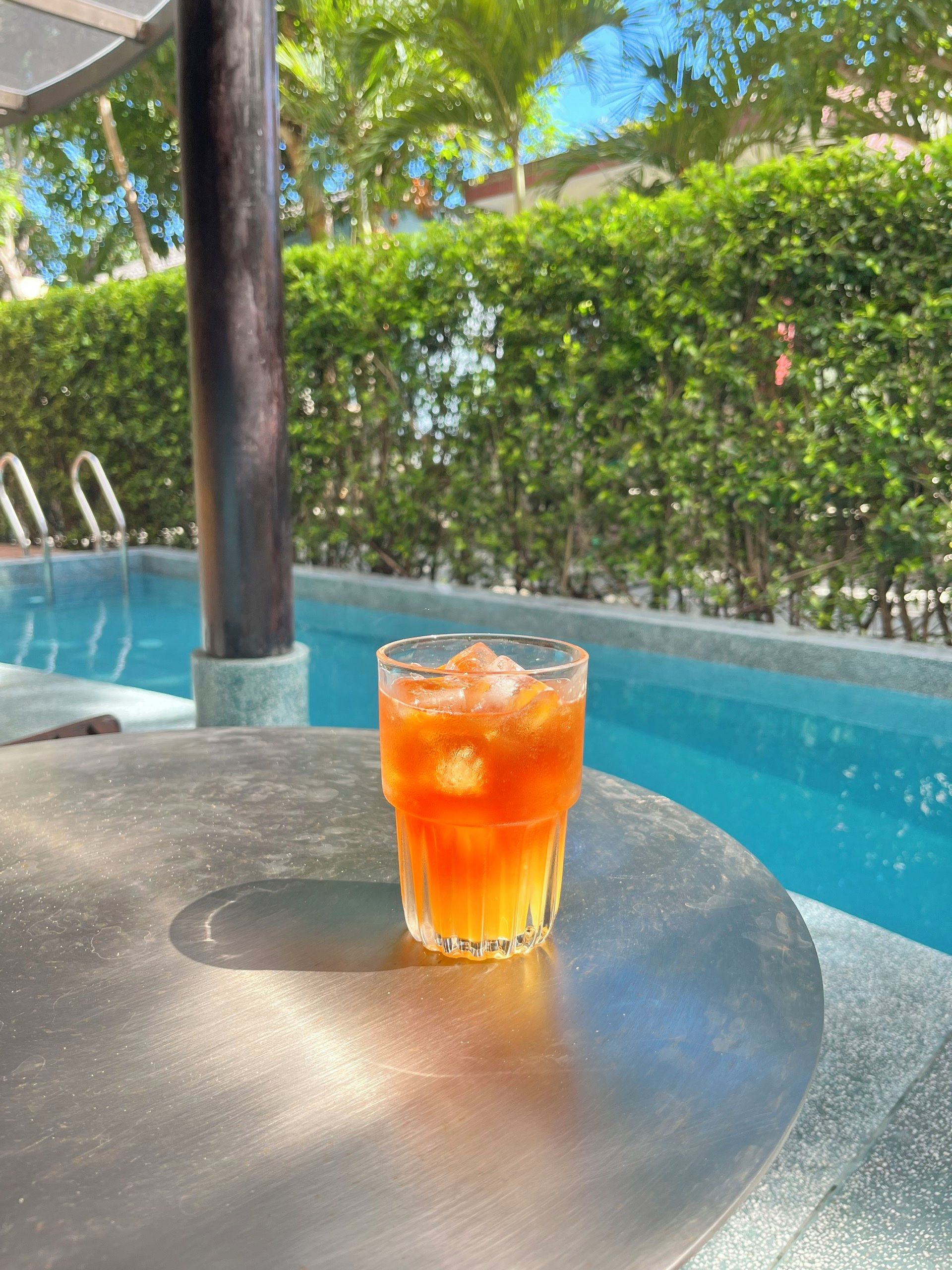 Ein Glas orangenes Getränk auf einem Metalltisch am Pool mit freiem Blick auf das Schwimmbecken und die umliegende Bepflanzung.