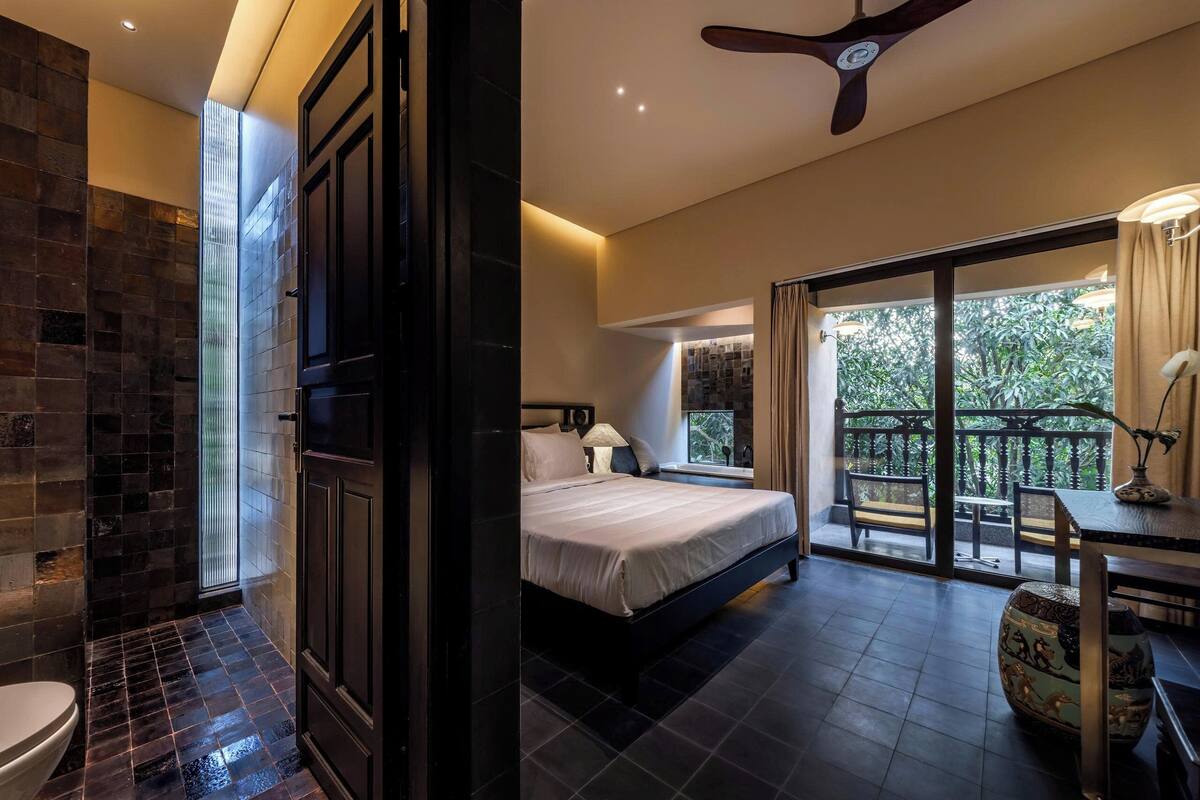 Un dormitorio con cama, mesita de noche, lámpara, florero y balcón con vistas al verdor.