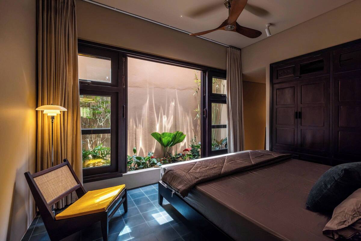 Un dormitorio con cama, una silla, ventana con cortinas, ventilador de techo y vista a zonas verdes.