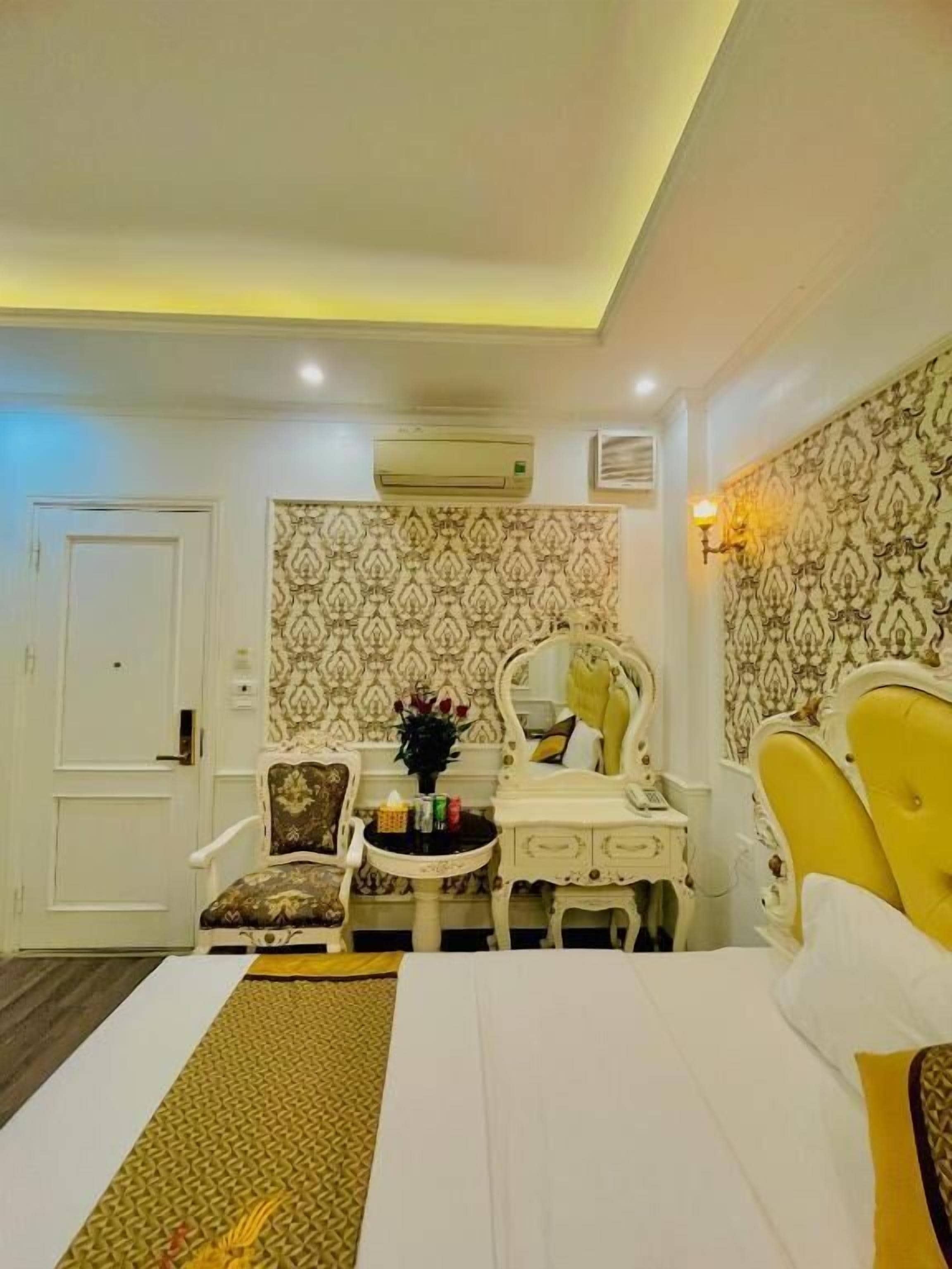 A hotel room with a bed, a vanity with a mirror, a small table with toiletries, patterned wallpaper, and an air conditioning unit.