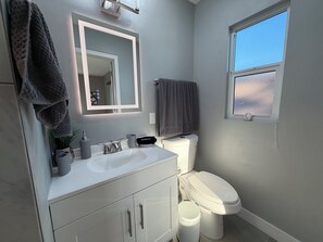Et badeværelse med hvid vask, spejl, toilet og et vindue.