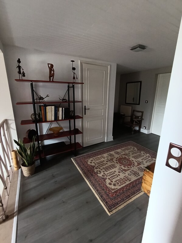 A hallway with a wooden floor, a patterned rug, a bookshelf with various items, a white door, and a mirror on the wall.