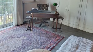 Una habitación con techo de madera, un ventanal con cortinas, un escritorio con una silla, un radiador, una lámpara de escritorio verde y una alfombra estampada.