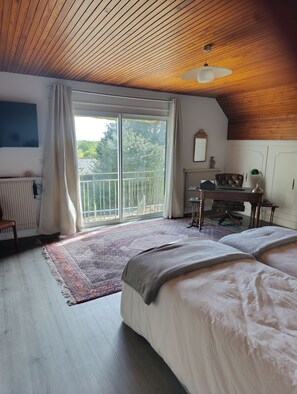 A bedroom with a large bed, a desk, a chair, a radiator, a window with curtains, and a ceiling with wooden beams.