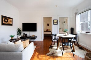 Ein modernes Wohnzimmer mit Flachbildfernseher, einer Essecke mit rundem Tisch und Stühlen, einer Couch und einem großen Fenster.