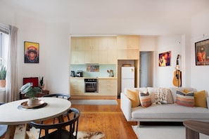 Eine moderne Küche mit Holzschränken, ein Essbereich mit rundem Tisch und Stühlen sowie ein Wohnzimmer mit Sofa und Kissen.