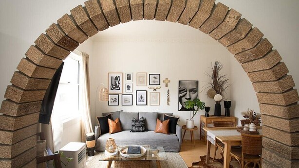 A cozy living room with a brick archway, a sofa with cushions, a coffee table, and various wall art. - Modern 2BR Boho Apt One Block From The Beach (Bondi Beach)
