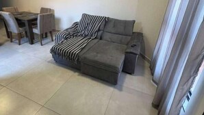 A living room with a grey sectional sofa, a striped throw blanket, a small table with chairs, and a window with curtains.
