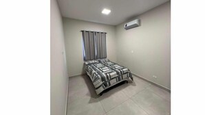 A bedroom with a bed, curtains, an air conditioning unit, and a tiled floor.