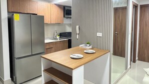 Uma cozinha compacta com geladeira de aço inoxidável, micro-ondas e fogão. Há uma pequena mesa de madeira com alguns pratos e um livro. A cozinha possui armários de madeira e painel de fundo cinza.