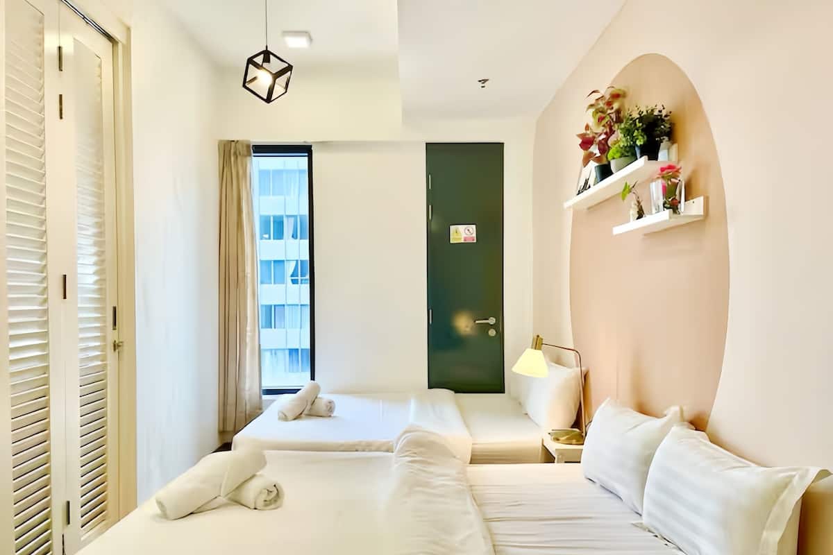 Ein Hotelzimmer mit zwei Betten, einem Fenster mit Vorhängen, einer grünen Tür, einem Regal mit Pflanzen und einer Pendelleuchte.