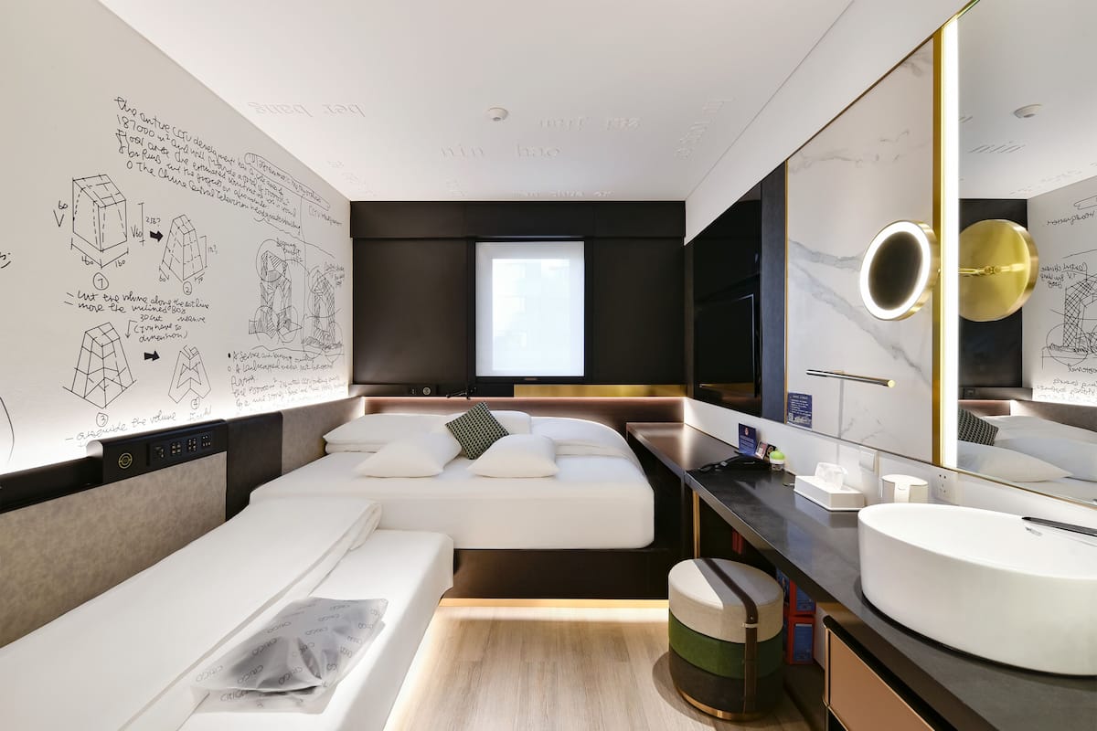 A modern hotel room with a large bed, a bedside table, a bathroom with a large mirror, and a window.