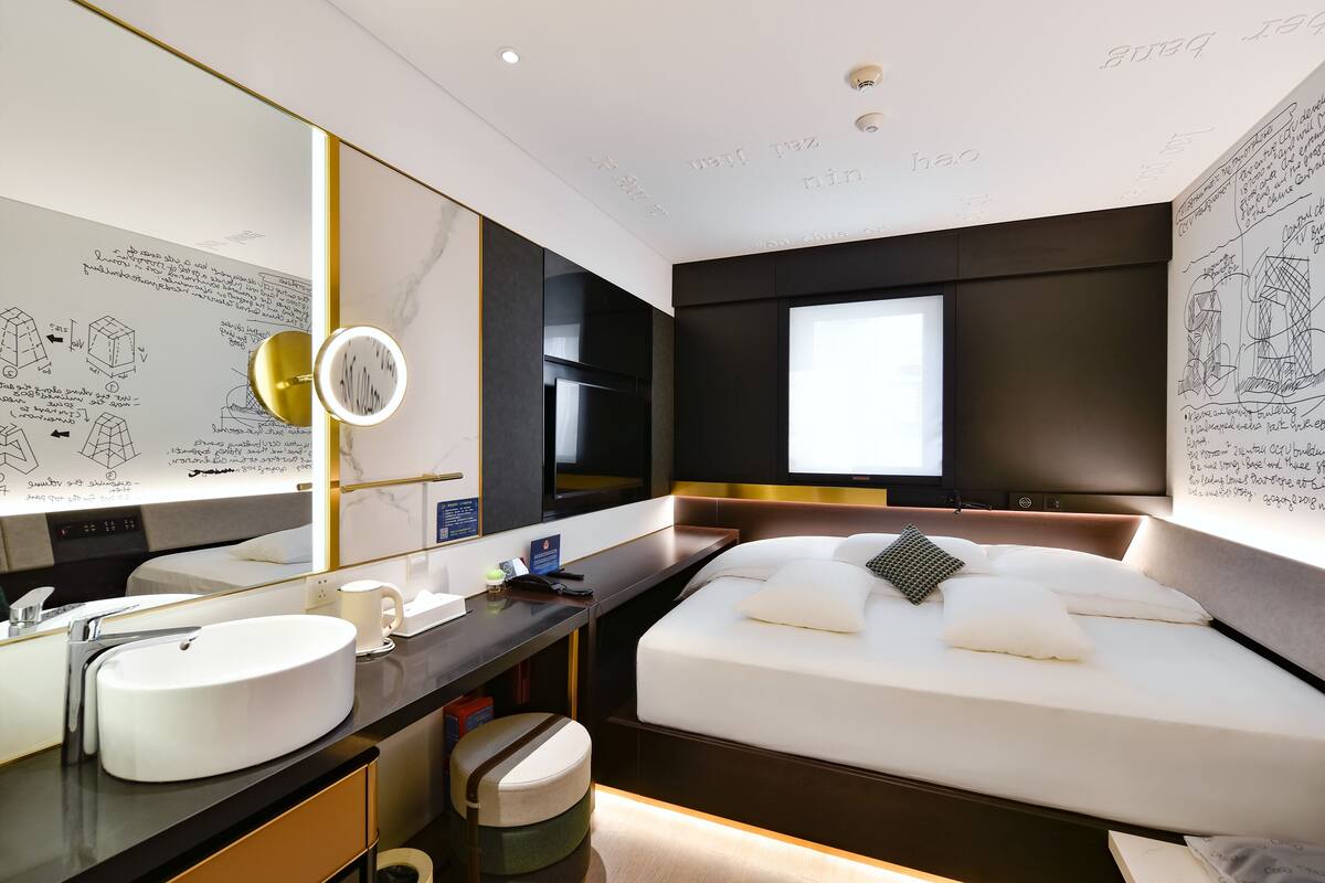 A modern hotel room with a large bed, a bedside table with a lamp, a large mirror, and a wall with architectural sketches.