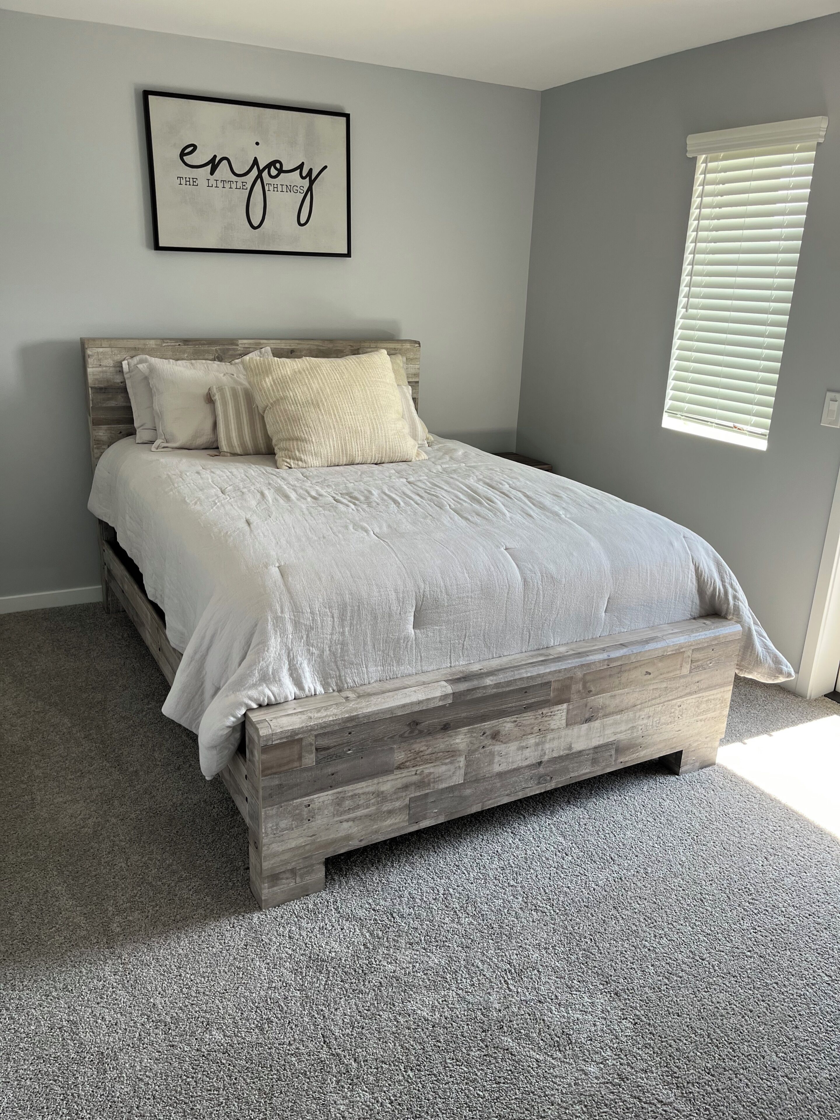 Een slaapkamer met een houten bedframe, een grijs tapijt, een grijze muur, een lijst met een citaat en een raam met jaloezieën.