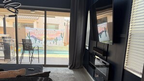 A modern living room with a large TV, dark furniture, and a view of a pool and building with a mural. - Luxury Glendale Stay with Pool & Theater Room by Cardinals Stadium (Glendale)