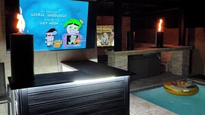 A pool area with a television displaying a cartoon, a ceiling fan, and a poolside seating area. - Luxury Glendale Stay with Pool & Theater Room by Cardinals Stadium (Glendale)